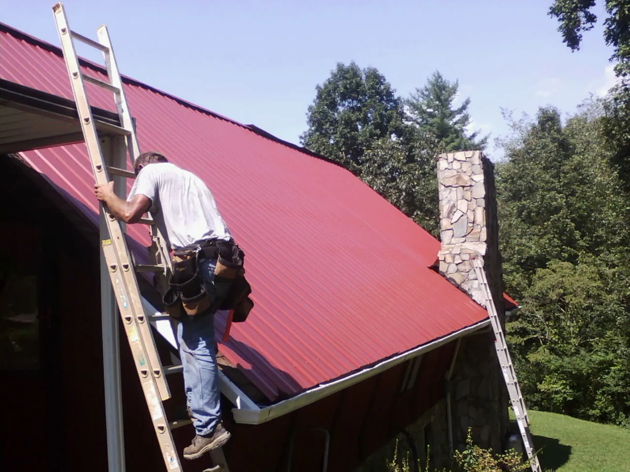 Roofer on a ladder beside a red metal roof and stone chimney — Soffit & Fascia Repair by Roofing Brothers in Monroeville